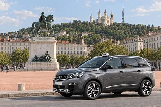 Berline grise avec enseigne taxi sur la place Bellecour, avec Fourvière visible en arrière-plan.